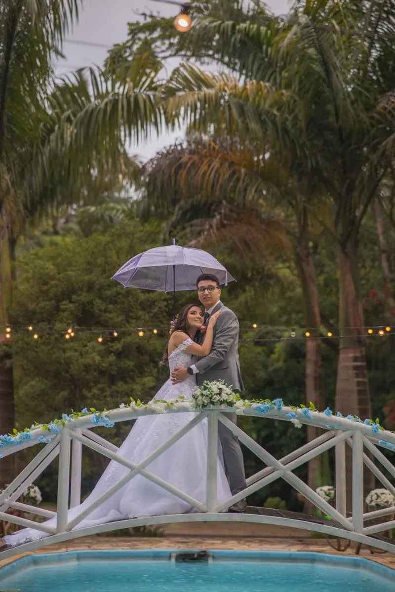 Casal em ensaio pré-wedding com guarda-chuva em jardim com piscina